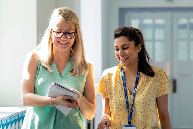 Two women walking indoors, smiling and talking. One holds a tablet and wears glasses, while the other wears a yellow shirt with a lanyard and badge—both discussing cancer prevention resources for health professionals in a sunlit corridor.