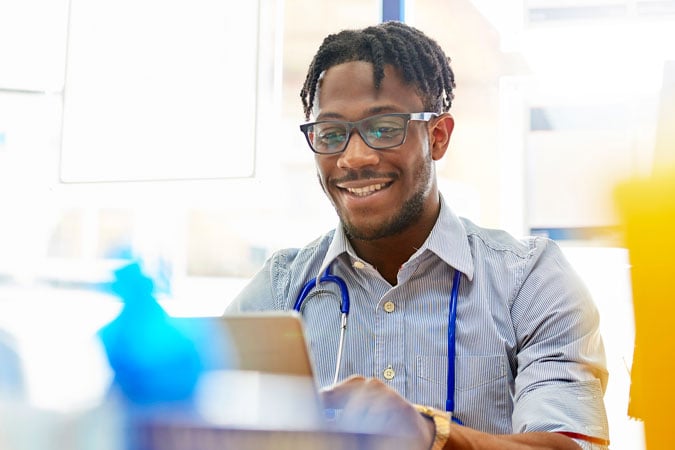 A smiling man wearing glasses and a stethoscope sits at a desk, typing on a laptop in a brightly lit room, searching for cancer prevention resources for health professionals.