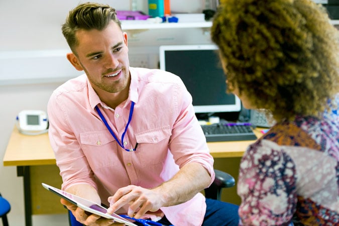 A male healthcare professional in a pink shirt discusses cancer prevention resources for health professionals on a tablet with a female patient in an office setting.