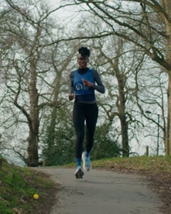A woman, Kemi Williams, is running outdoors on a paved path surrounded by bare trees. Wearing glasses and a blue vest, she trains for the London Marathon with a focused expression, inspired by her cancer fundraising story.