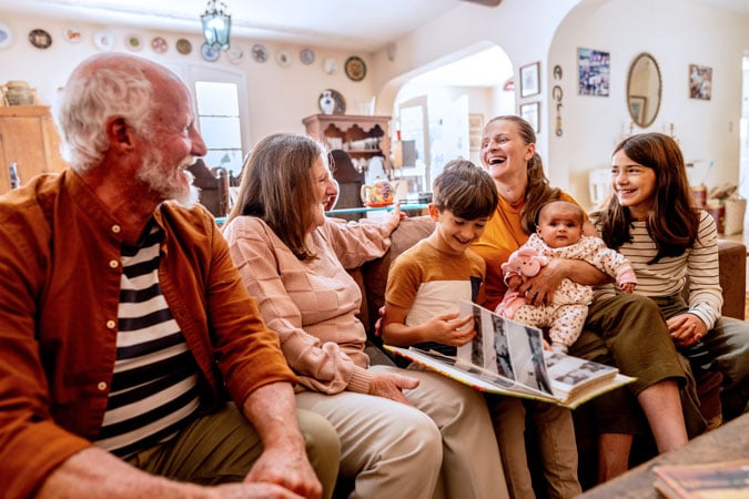 Three generations of a family sit together on a sofa, smiling and laughing. An older couple, two women, a young boy holding a photo album, and a baby are gathered in a cosy, warmly lit sitting room.
