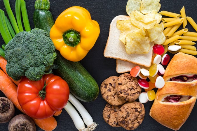 An assortment of fresh vegetables sits beside ultra-processed foods (UPFs) like crisps and biscuits, highlighting the contrast between wholesome choices and items increasingly linked to health concerns such as cancer.