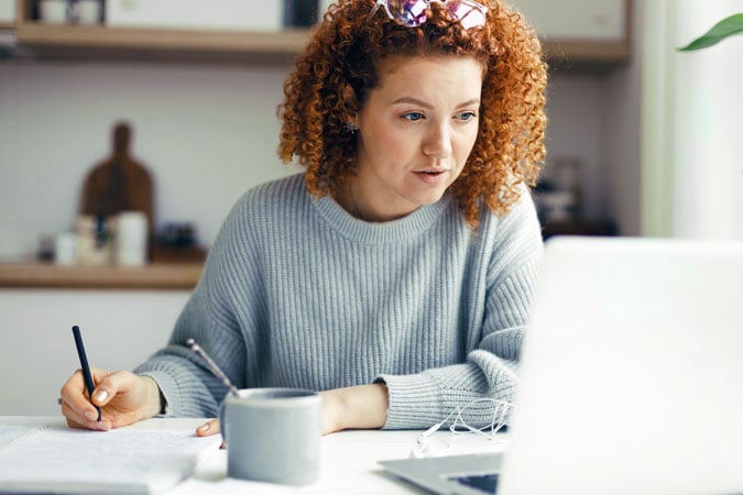 A woman with curly red hair in a grey jumper sits at a table, taking notes in a notebook whilst looking at cancer prevention learning resources for health professionals on her laptop. A mug, papers, and spectacles are also on the table.