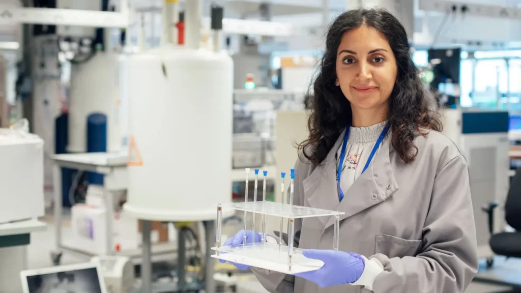 A woman in a lab coat and gloves is standing in a laboratory, smiling at the camera and holding a tray with pipettes. Laboratory equipment and shelves are visible in the background.
