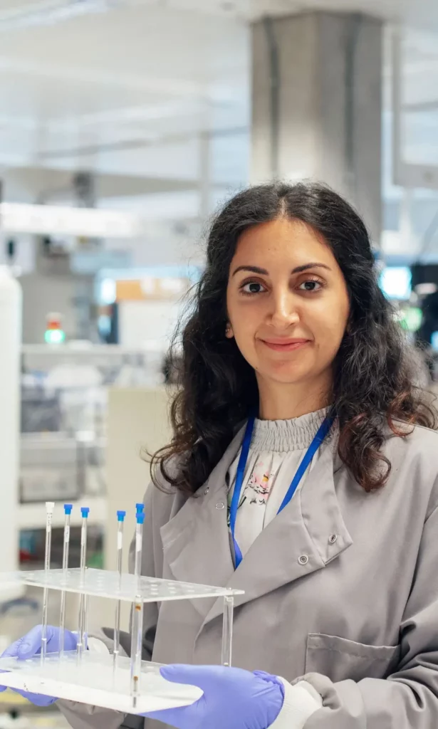 A woman in a lab coat and gloves is standing in a laboratory, smiling at the camera and holding a tray with pipettes. Laboratory equipment and shelves are visible in the background.