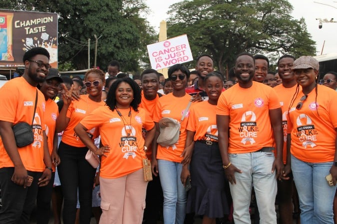 A group of people wearing bright orange shirts stand together outdoors, smiling at the camera. One person holds a sign that reads CANCER IS JUST A WORD, raising awareness about breast cancer in sub-Saharan Africa. Trees and a billboard are visible in the background.