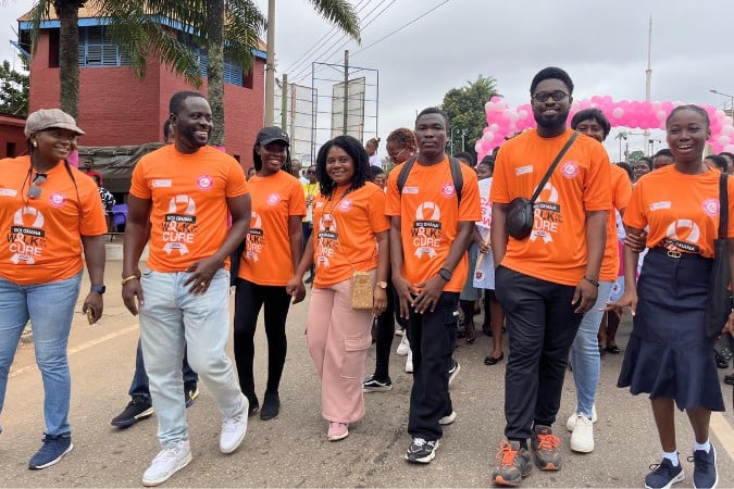 A group of people wearing matching orange T-shirts with a breast cancer awareness message walk together outdoors, smiling and participating in an organised event highlighting breast cancer in sub-Saharan Africa. Palm trees and a building are visible in the background.