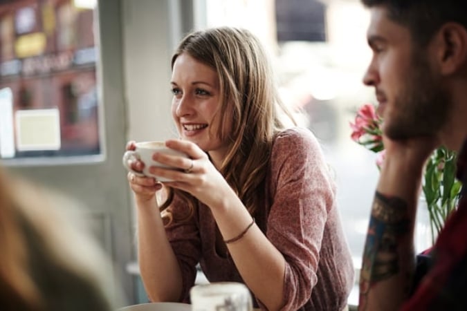A woman smiling and holding a cup while sitting at a table in a cafe, with a man seated beside her. Sunlight comes through the window, and the atmosphere appears relaxed and casual.