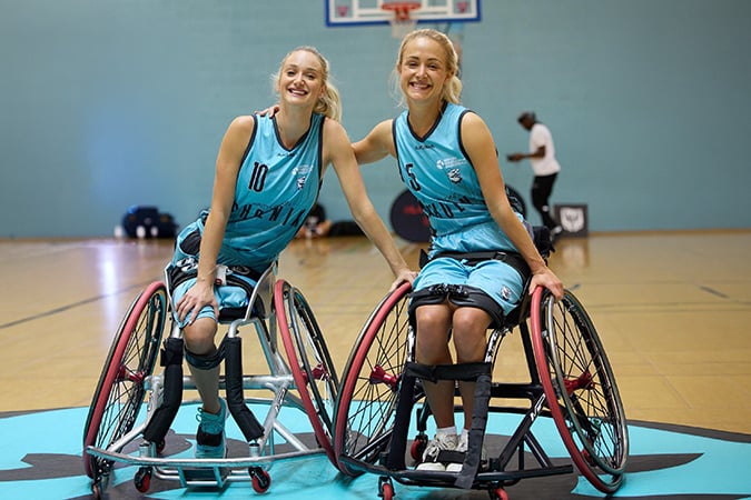 Two young women in teal sports kits sit in wheelchairs on an indoor basketball court, smiling and posing for the camera. A basketball net and another person are visible in the background.