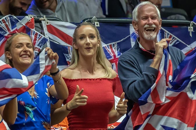 Three people smile and cheer whilst holding British flags, with Union Jack bunting in the background. The group appears joyful and festive, suggesting they are celebrating or supporting a UK-related event.