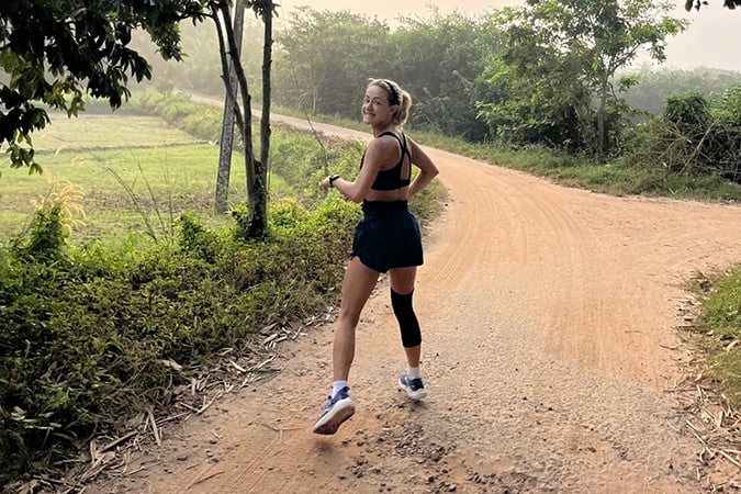 A woman in sportswear runs on a dirt road surrounded by greenery, looking back and smiling on a misty morning. She wears a knee support and white trainers.