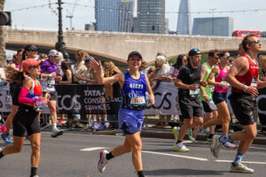 Runners participate in a city marathon, with one smiling runner in a blue outfit waving at the camera. Crowds cheer from behind barriers, and tall buildings are visible in the background.
