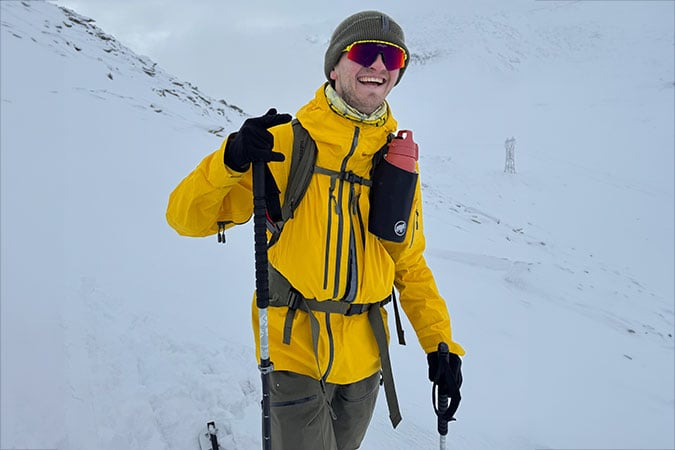 A ski instructor wearing a yellow jacket, green beanie, and red sunglasses smiles whilst standing on a snowy mountain slope with walking poles and a water bottle attached to their rucksack, undeterred by their rare cancer diagnosis.