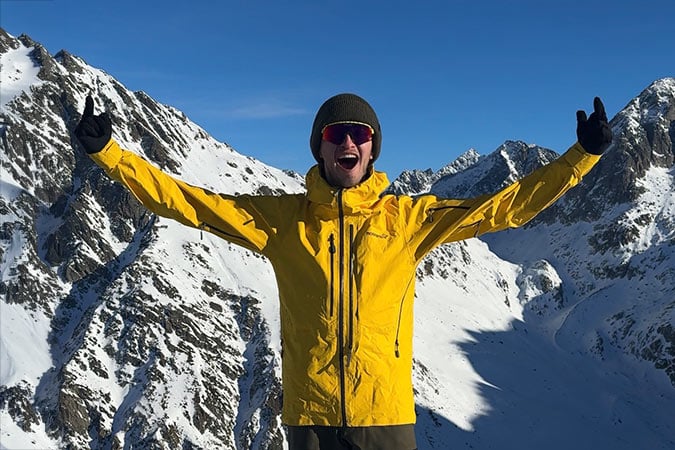 A ski instructor in a yellow jacket and sunglasses stands on a snowy mountain with arms raised, smiling against a clear blue sky. Snow-covered peaks fill the background, celebrating life after a rare cancer diagnosis.