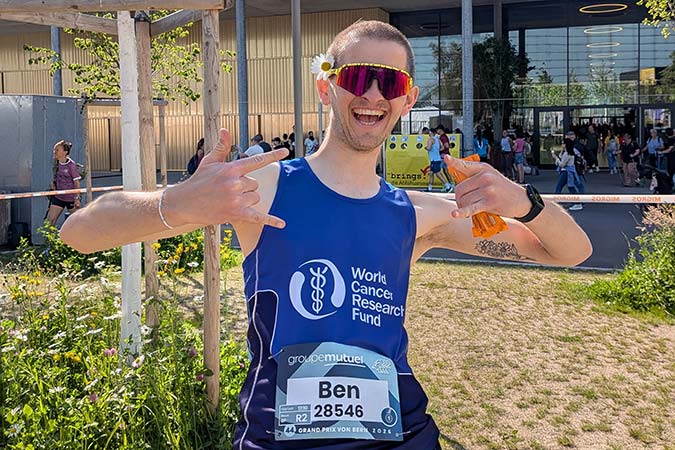A smiling runner, a ski instructor who overcame a rare cancer diagnosis, wears a blue World Cancer Research Fund vest and race number labelled Ben 28546, making hand signs and posing outdoors near a building with people and greenery in the background.