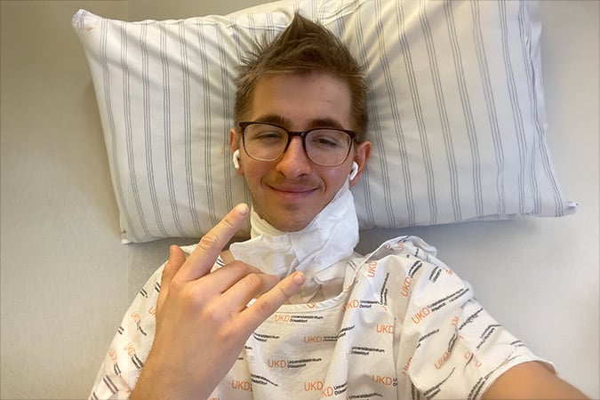 A young person with glasses is lying on a bed in a hospital gown and neck bandage, smiling despite a rare cancer diagnosis. Wireless earphones in, they make a peace sign; behind them rests a striped pillow, hinting at their former life as a ski instructor.