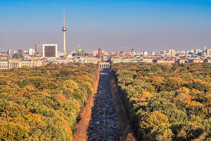 Aerial view of Berlin with the Brandenburg Gate in the centre, a wide avenue lined with autumn trees, and the TV Tower rising above city buildings in the background.