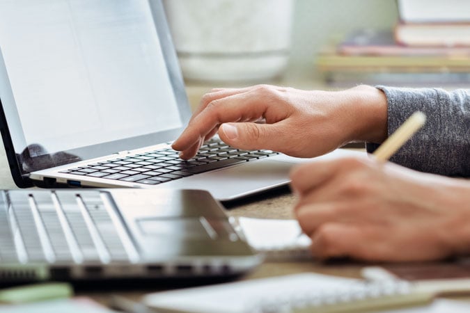 A person types on a laptop with one hand while writing notes with a pencil in the other hand. Another laptop and some notebooks are visible on the desk.