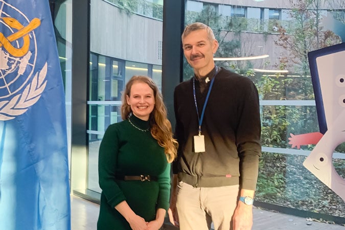 Two people stand indoors, smiling at the camera. A blue flag with a yellow symbol is on the left, and large windows reveal plants and a modern building outside—perhaps at an event discussing the new definition of obesity beyond BMI. The right person wears a lanyard and ID badge.