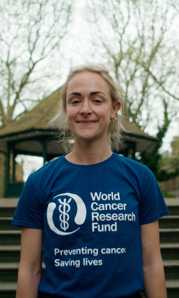 A woman stands outdoors, smiling, wearing a blue World Cancer Research Fund T-shirt that reads Preventing cancer. Saving lives. Trees and a gazebo appear in the blurred background.