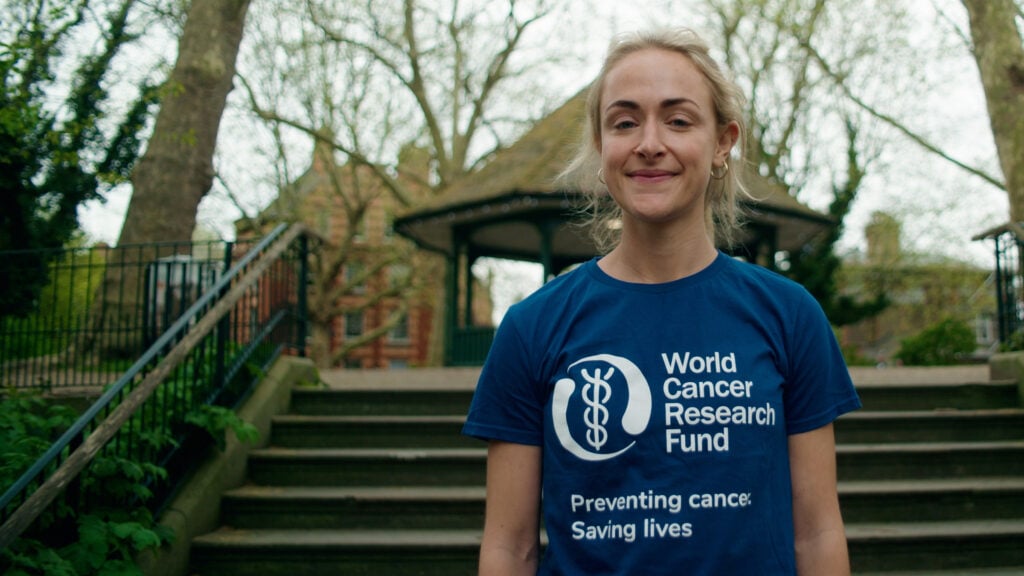 A woman stands outdoors in front of stone steps and a bandstand, smiling and wearing a blue World Cancer Research Fund T-shirt that says Preventing cancer. Saving lives. Trees without leaves are visible in the background.