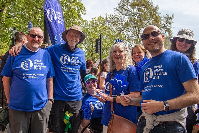 A group of smiling people wearing blue World Cancer Research Fund shirts gather outdoors at an event, with trees in the background and others in the crowd behind them.