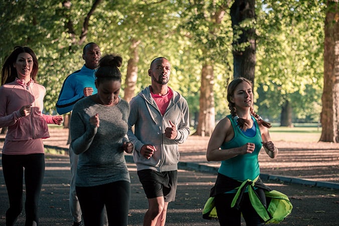 A group of five adults jog together along a shaded park path, participating in cancer fundraising events. Surrounded by green trees and sunlight, they appear focused and are dressed in sports clothing.