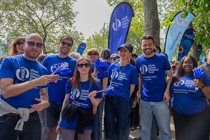 A group of people wearing blue World Cancer Research Fund T-shirts smile and pose together outdoors, holding blue clackers. Blue banners and trees are visible in the background.