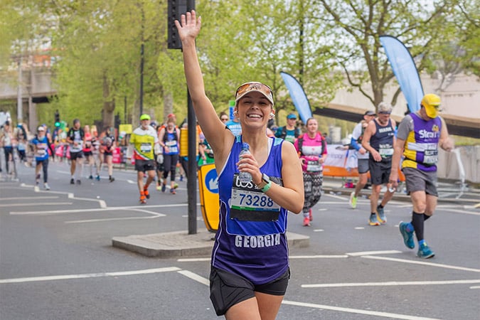 A smiling woman runner waves at the camera while holding a water bottle during a cancer fundraising event marathon. Other participants run behind her on a city street lined with trees and banners.