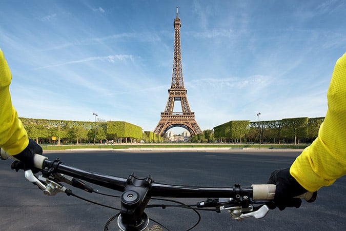 View from a cyclist's perspective of the Eiffel Tower, with hands on the handlebars, wearing yellow sleeves, approaching the famous Paris landmark on a clear day with green trees lining the path.