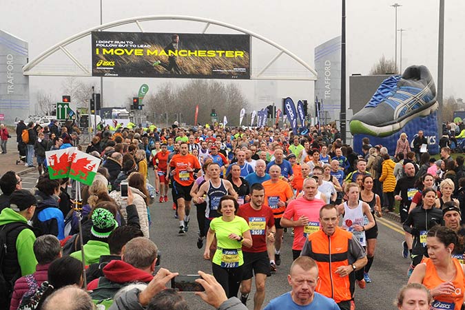 A large crowd of marathon runners in colourful outfits race past cheering spectators near a start line with a sign reading I MOVE MANCHESTER. The scene is lively, with banners and a giant trainer on display.