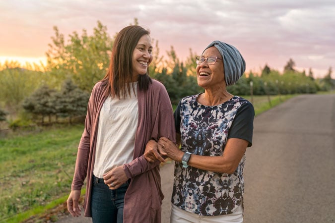 Two women, one younger and one older, walk arm in arm down a paved path outdoors at sunset, smiling and looking at each other, with trees and greenery in the background.
