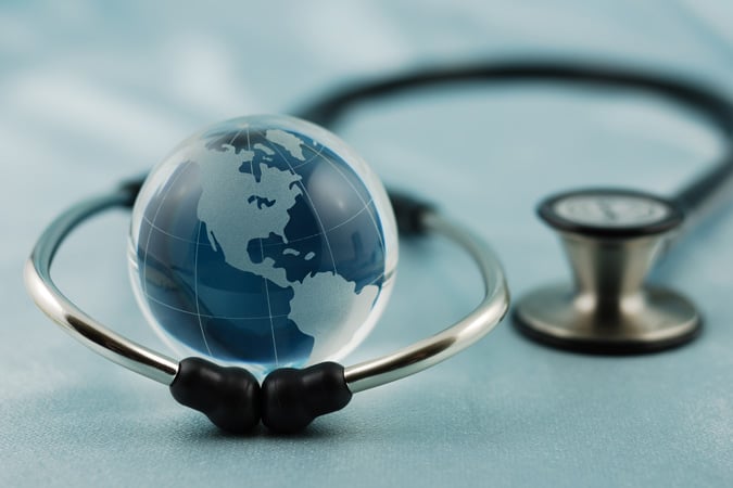 A glass globe with a map of the Americas sits inside the circular part of a stethoscope, symbolising global health or international cancer control policy, on a soft blue surface.