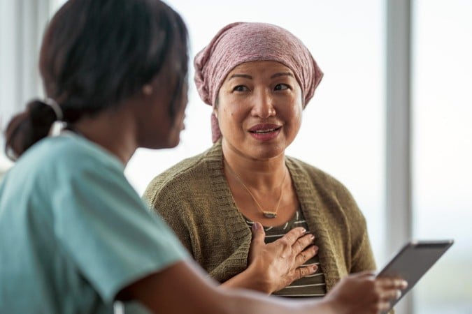 A woman wearing a headscarf and cardigan sits with her hand on her chest, discussing cancer control policy with a healthcare professional holding a tablet in a medical setting.