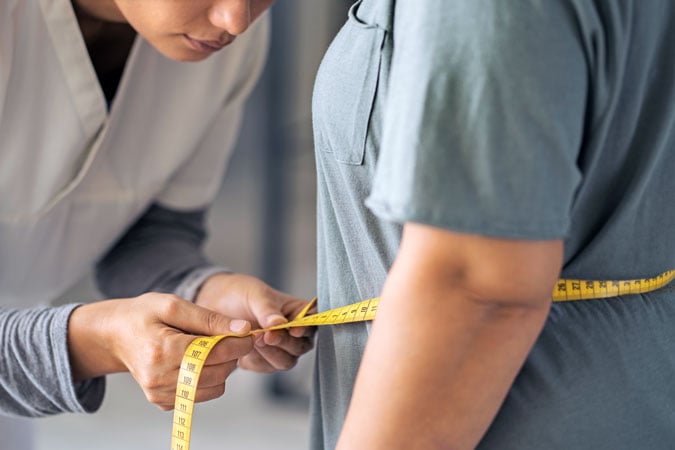 A healthcare professional uses a yellow measuring tape to assess waist size, reflecting the new definition of obesity beyond BMI, whilst only their upper bodies and hands are visible.
