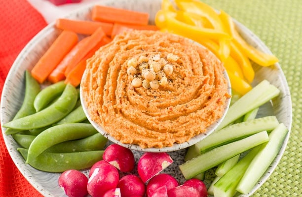 A plate with a bowl of houmous topped with chickpeas and pepper, surrounded by fresh vegetables: carrot sticks, yellow pepper strips, cucumber sticks, sugar snap peas, and radishes.