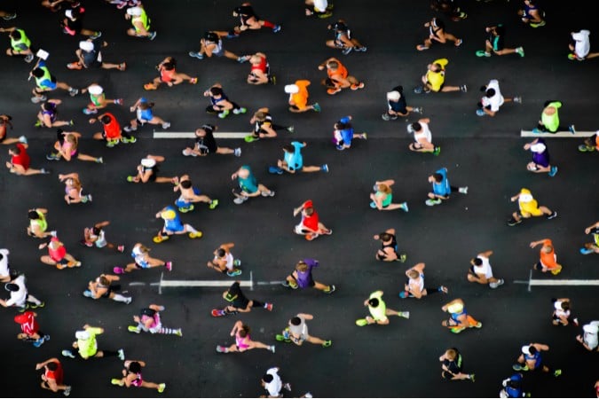Aerial view of numerous runners in colourful athletic clothing participating in a marathon on a dark tarmacked road, captured from above. The runners are spaced out and moving in the same direction.