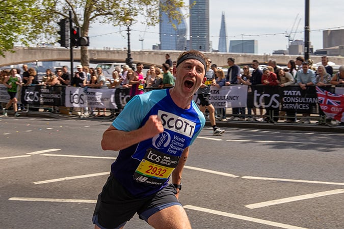 A male runner in a blue shirt labelled Scott with race number 2932 enthusiastically sprints past cheering spectators during a city marathon, with skyscrapers and the Shard visible in the background.
