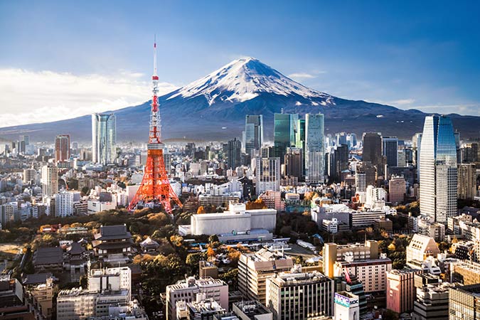 A cityscape of Tokyo with modern skyscrapers, Tokyo Tower in the centre, and snow-capped Mount Fuji in the background under a clear blue sky.