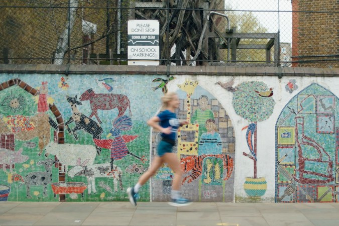 A person in blue sportswear jogs past a colourful mosaic mural of animals, trees, and people—perhaps inspired by local cancer fundraising events. A sign above reads PLEASE DON'T STOP ON SCHOOL MARKINGS.