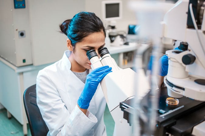 A scientist wearing a white lab coat and blue gloves looks through a microscope in a laboratory, researching how blocking fat production could slow breast cancer metastasis, with scientific equipment visible in the background.