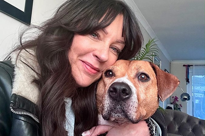 A woman with long dark hair smiles while hugging a brown and white dog. They are indoors on a sofa, with a plant and a window visible in the background.