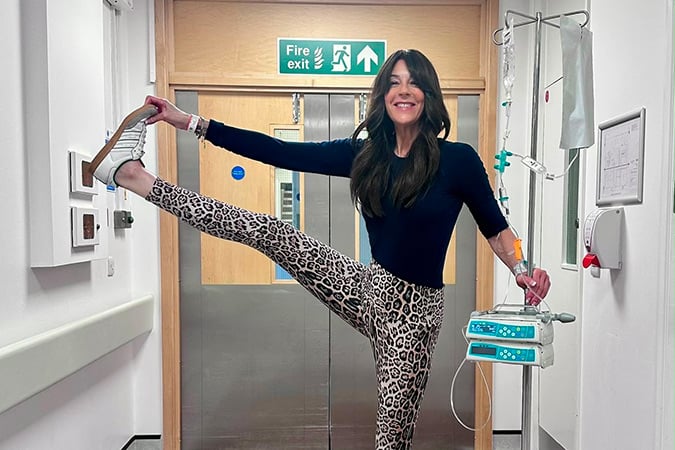 A woman in leopard-print leggings and a black top stretches her leg high against a hospital wall while smiling. She is attached to a drip and standing in a corridor, with a fire exit sign visible above the door behind her.