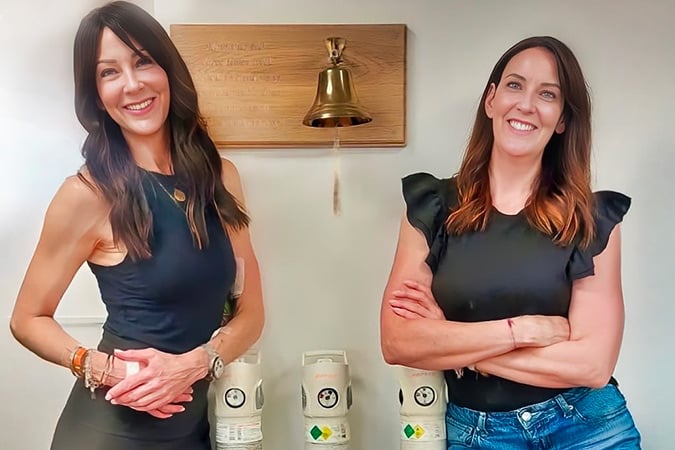 Two women with long brown hair stand smiling in front of a wall-mounted bell, with their arms folded. Three oxygen cylinders are visible on the floor behind them.
