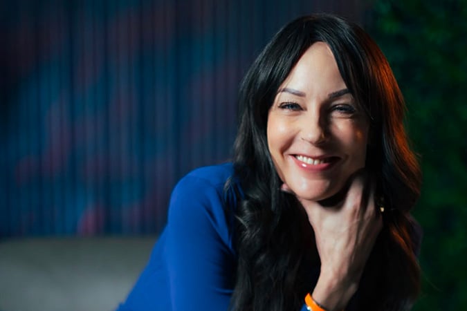 A woman with long dark hair, wearing a blue top, smiles warmly at the camera with her hand gently resting under her chin. The background is softly blurred with hints of blue and green.