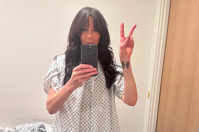 A person with long dark hair, wearing a patterned hospital gown, takes a mirror selfie and makes a peace sign with their left hand. The background shows a plain wall and an open wooden door.
