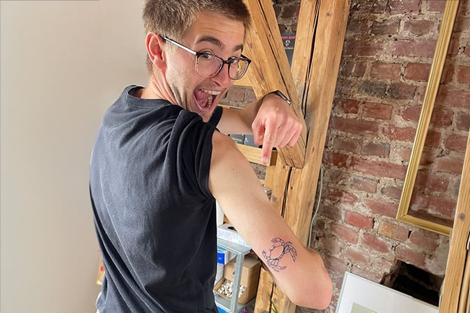 A smiling person wearing glasses pulls up their sleeve and points excitedly to a fresh crab tattoo on their upper arm, celebrating overcoming a rare cancer diagnosis, standing indoors near a brick wall and wooden beams.