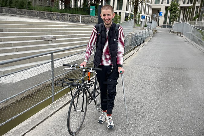 A person with a leg brace and crutch, smiling despite a rare cancer diagnosis, stands outdoors on a paved path holding a bicycle. Steps and buildings are visible in the background, reflecting the resilience of this former ski instructor.