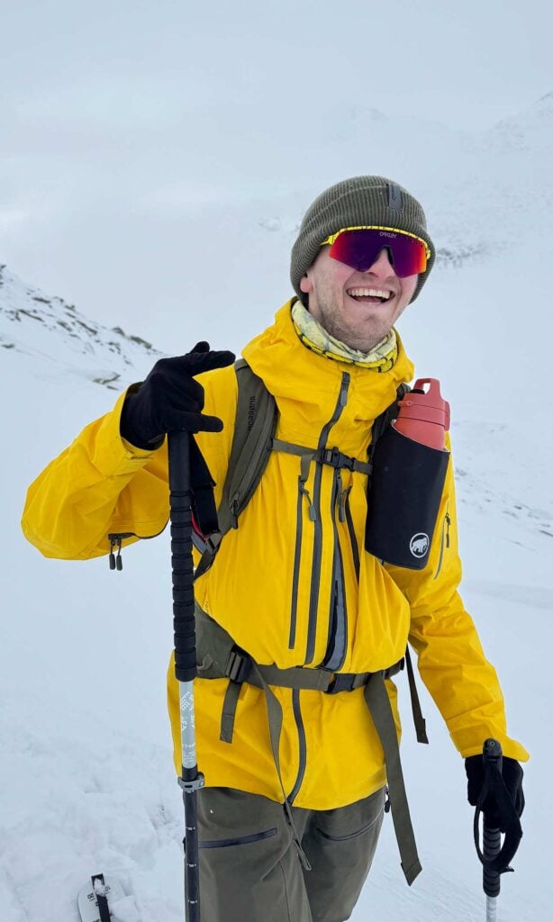 A person in a yellow jacket and grey beanie, wearing reflective sunglasses, stands on a snowy mountain slope holding a trekking pole and smiling. A red water bottle is attached to their rucksack.
