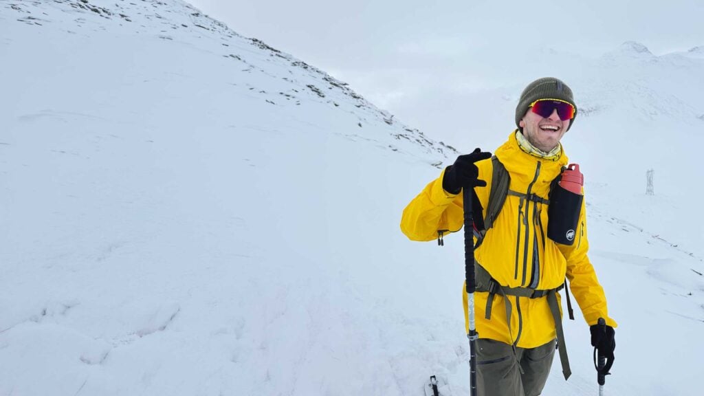 A person wearing a yellow jacket, green beanie, sunglasses, and gloves smiles whilst holding walking poles on a snowy mountain slope. A red water bottle hangs from their rucksack. Snow-covered peaks are in the background.
